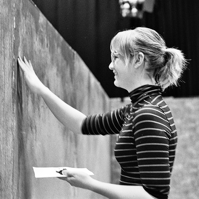 A black and white film photo of a young white woman holding a palette and using her remaining hand to paint walls of a film set.