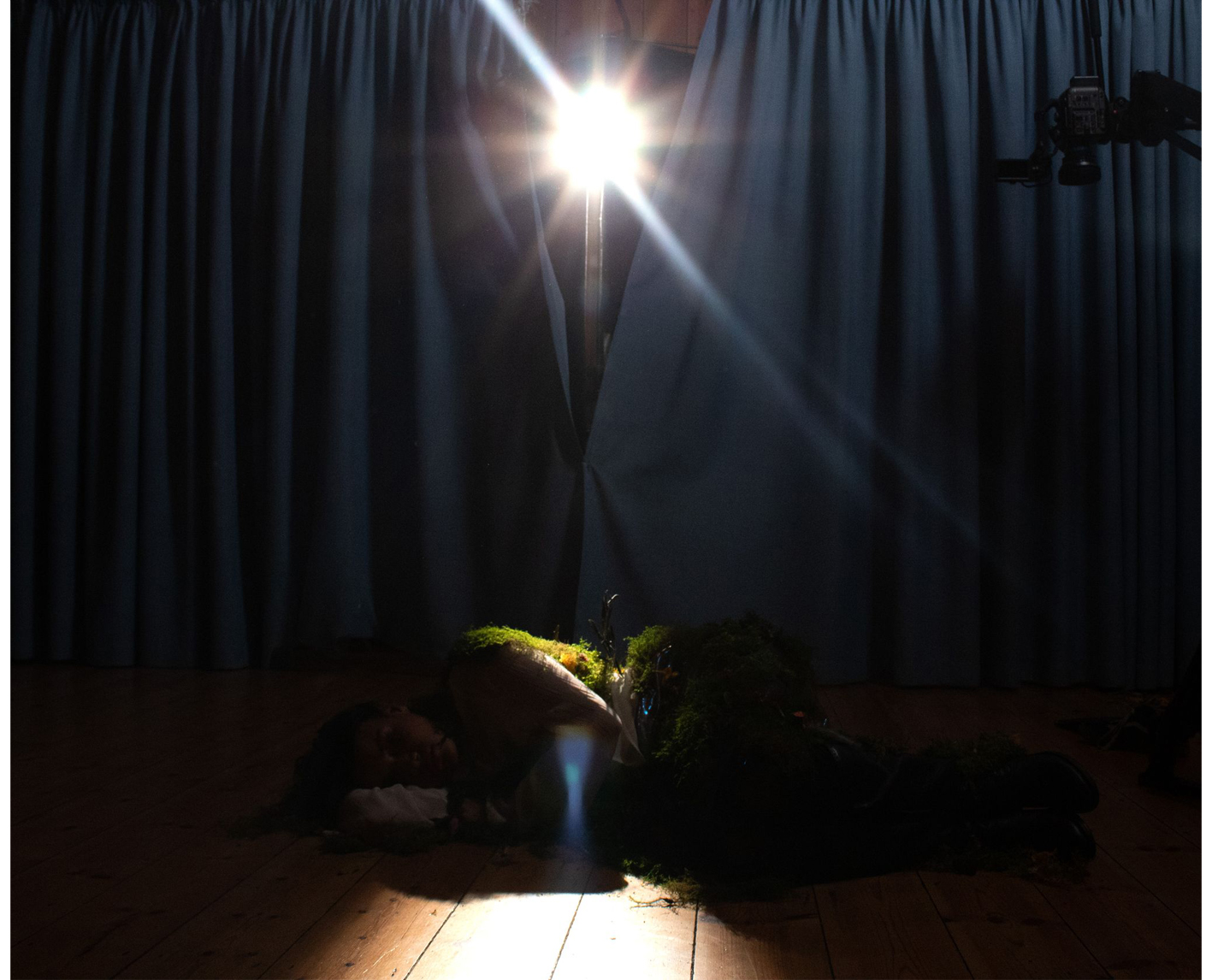 Girl lying on a stage floor covered in moss with bright light shining through stage curtains
