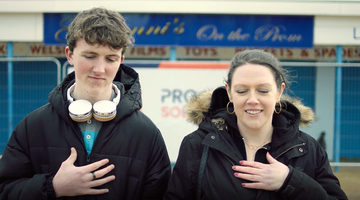 Ellis Kavanagh as Aaron and Laura Ollerton as Marie, stand with their hands on their chests on the promenade on Barry Island. Film still from What I Meant to Say.